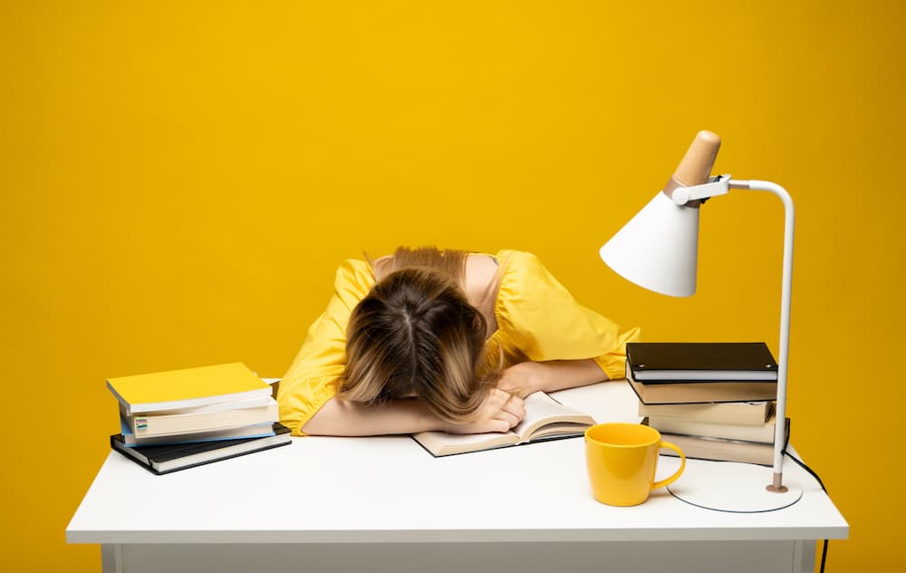 Woman resting her head on a desk surrounded by books and a coffee mug, symbolizing semaglutide fatigue and low energy during GLP-1 therapy.