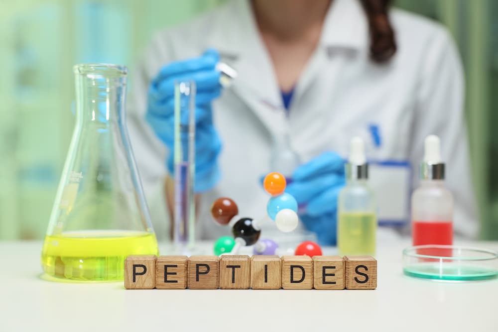 Lab setup showing a scientist with test tubes and the word “Peptides” spelled in wooden blocks, symbolizing peptide therapy and research.