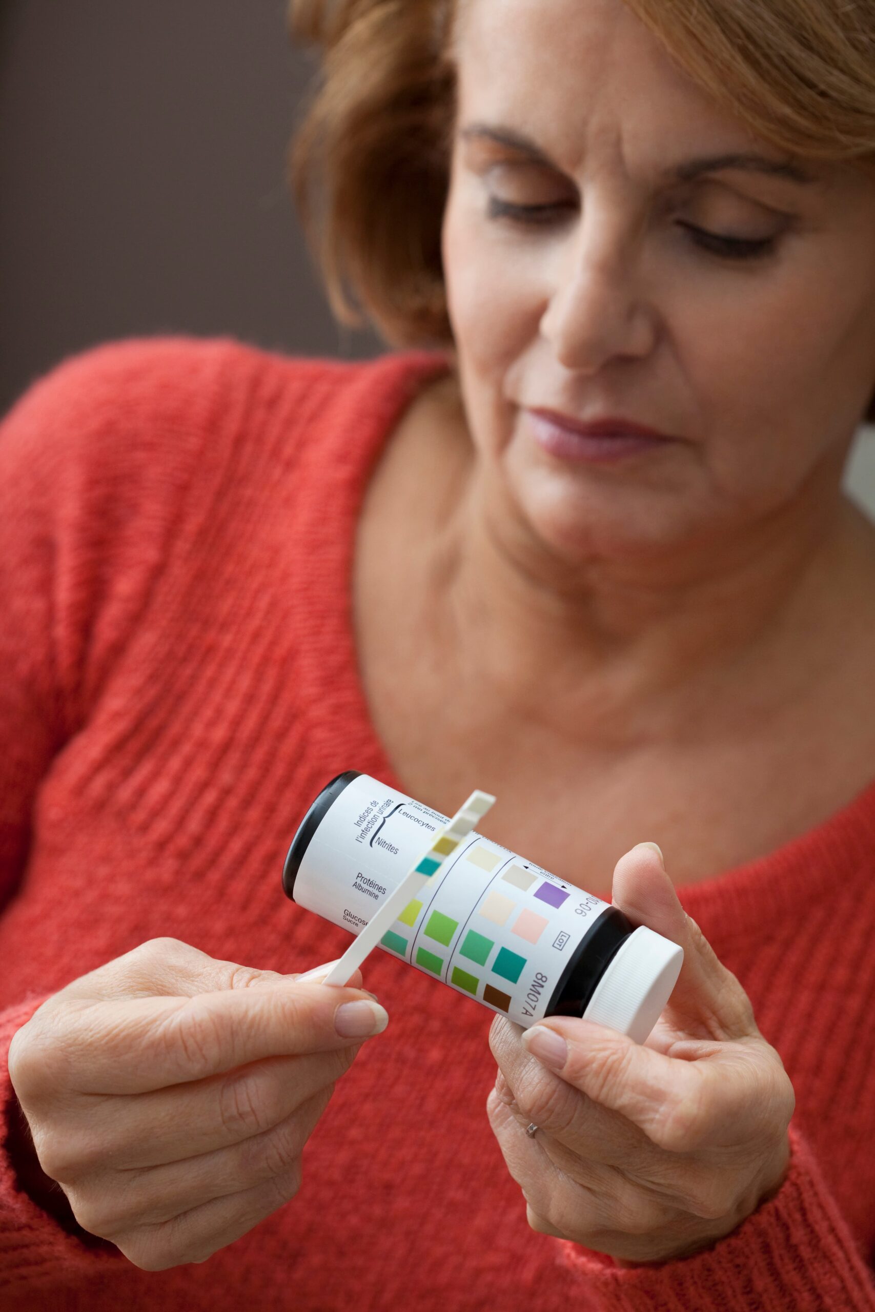 A concerned middle-aged woman in a red sweater examining a home urine test strip to check for recurrent UTI indicators like leukocytes and nitrites