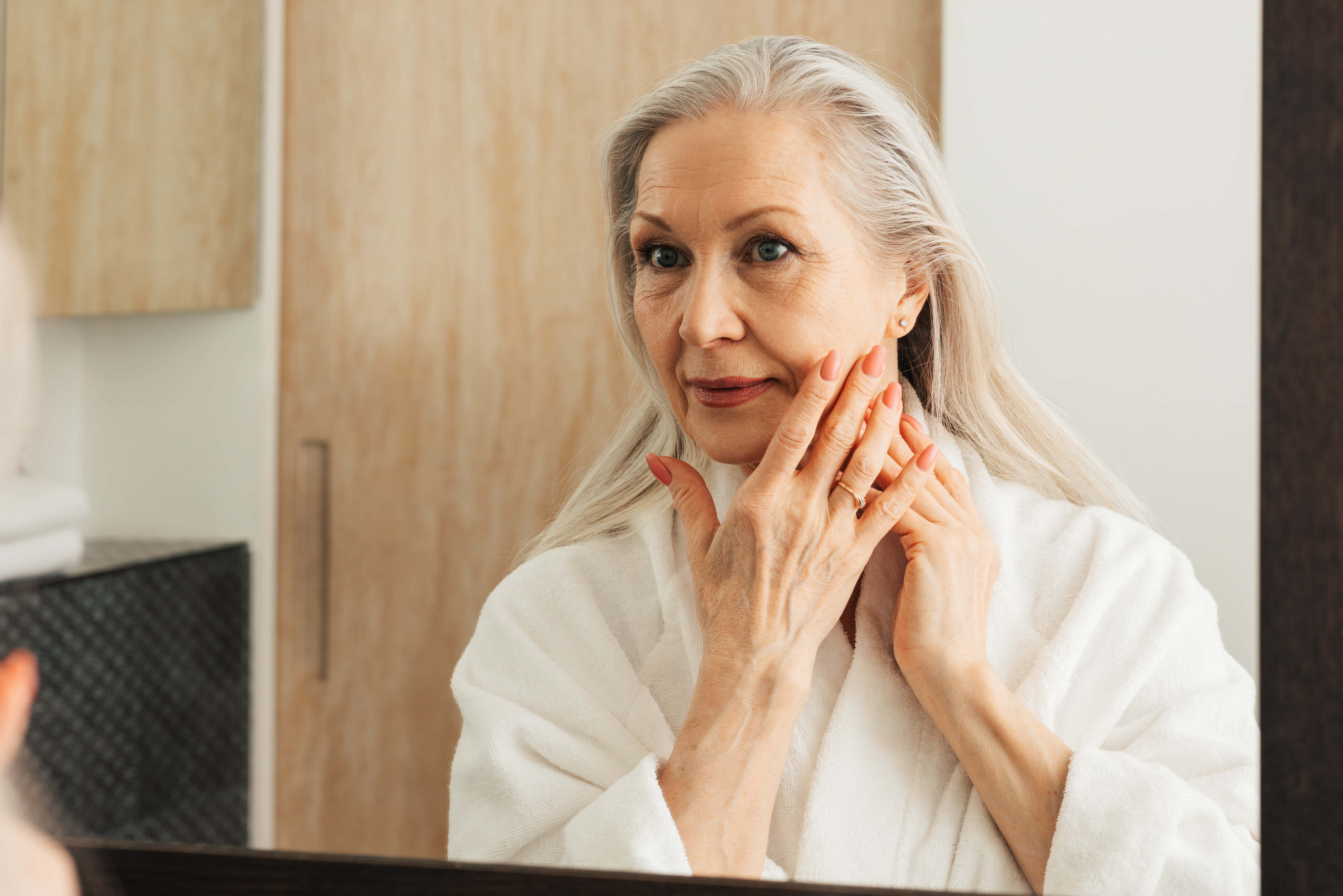 Mature woman with long silver hair in a white robe gently lifting and examining sagging jowls and loose jawline skin in the mirror, considering non-surgical Morpheus8 treatment.